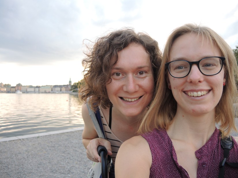 Foto van Sophie en Tamar op vakantie in Stockholm. Tamar zit in haar rolstoel, waarvan alleen één duwhendel te zien is. Ze heeft goudblond haar, grote donkerblauwe bril, en een zomers bordeaurood hemdbloesje met kleine witte stipjes aan. Ze staat lachend op de foto. Sophie staat achter haar en bukt zodat haar lachende gezicht naast dat van Tamar komt. Sophie heeft bruine kruillen, grote bruine ogen en draagt een beige hemdje met zwarte strepen. Op de achtergrond zondondergang over Stockholm.